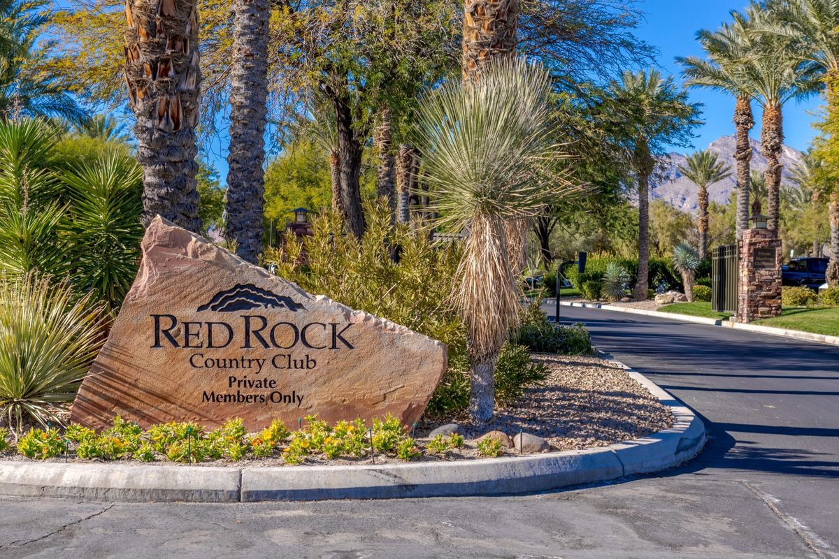 A carved sandstone boulder sign reading Red Rock Country Club, Private Members Only, with palm trees, yucca and flowers along the curb, a paved entry road with stone pillars on the right, and a mountain peak behind, in Summerlin, Las Vegas, Nevada.